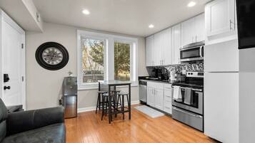 A modern kitchen with white cabinets, stainless steel appliances, a dining area with stools, and a large window with blinds.