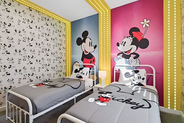 A children's room with two beds, each featuring Mickey Mouse designs. The room has a patterned wallpaper and a vibrant pink accent wall.
