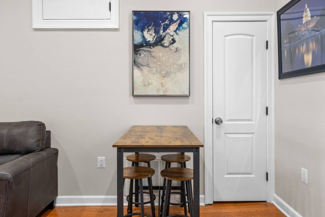 A room with a gray wall, a white door, a brown table with stools, and a gray couch.