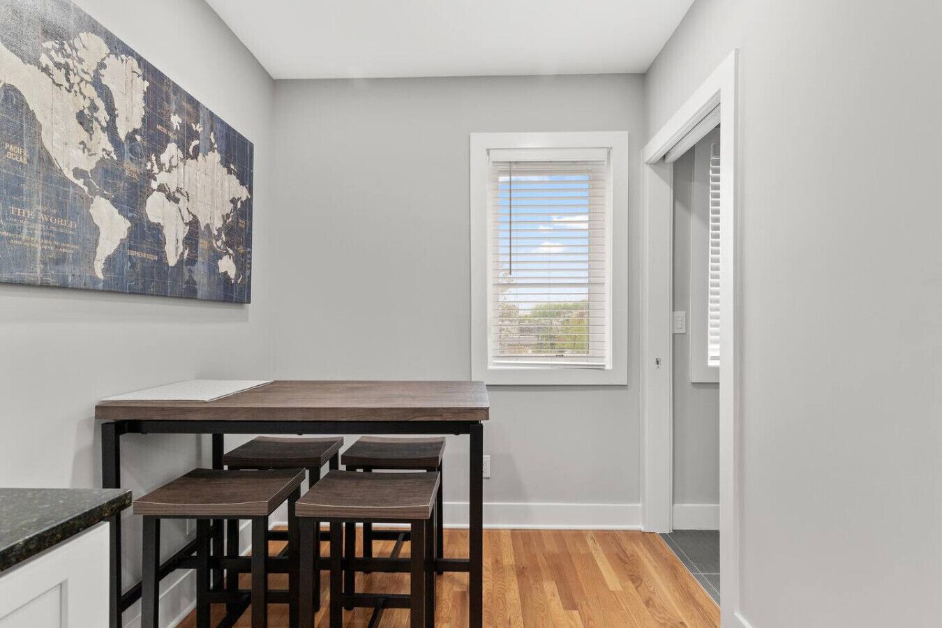 A kitchen with a wooden table and stools, a world map on the wall, and a window with blinds.