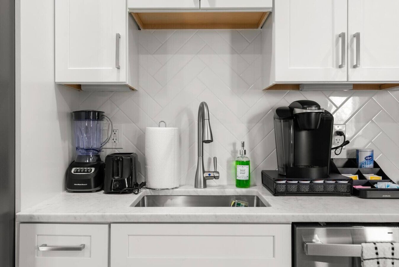 Eine moderne Küche mit weißen Schränken, einer Edelstahlspüle, einer Kaffeemaschine, einem Mixer, einem Toaster und einem Spülmittelspender.