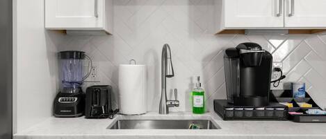 Eine moderne Küche mit weißen Schränken, einer Edelstahlspüle, einer Kaffeemaschine, einem Mixer, einem Toaster und einem Spülmittelspender.