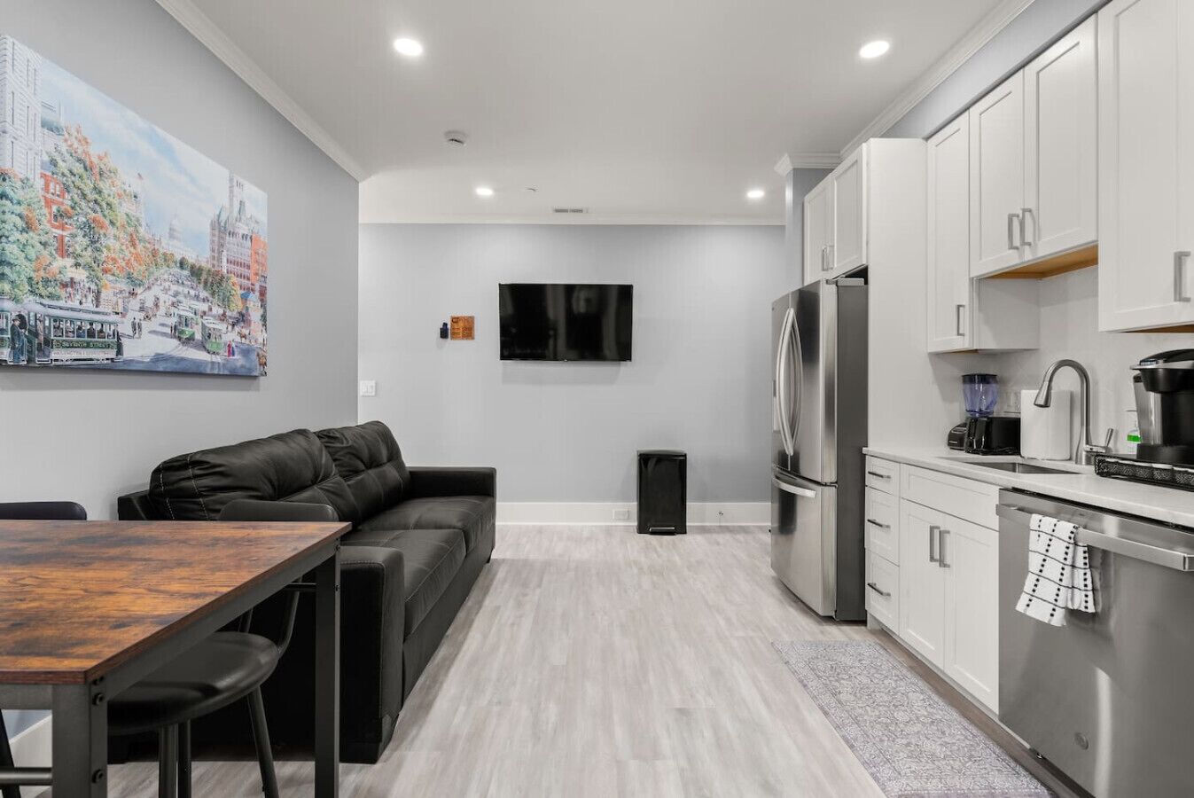 A modern kitchen with a built-in oven, a refrigerator, and a coffee maker. There is a black leather sofa and a dining table with chairs.