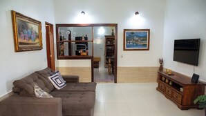 A living room with a brown sectional sofa, a wooden TV stand, and a large flat-screen TV. There is a painting on the wall and a door leading to another room. - Casa com Piscina e Churrasqueira no Pantanal (Angra dos Reis)