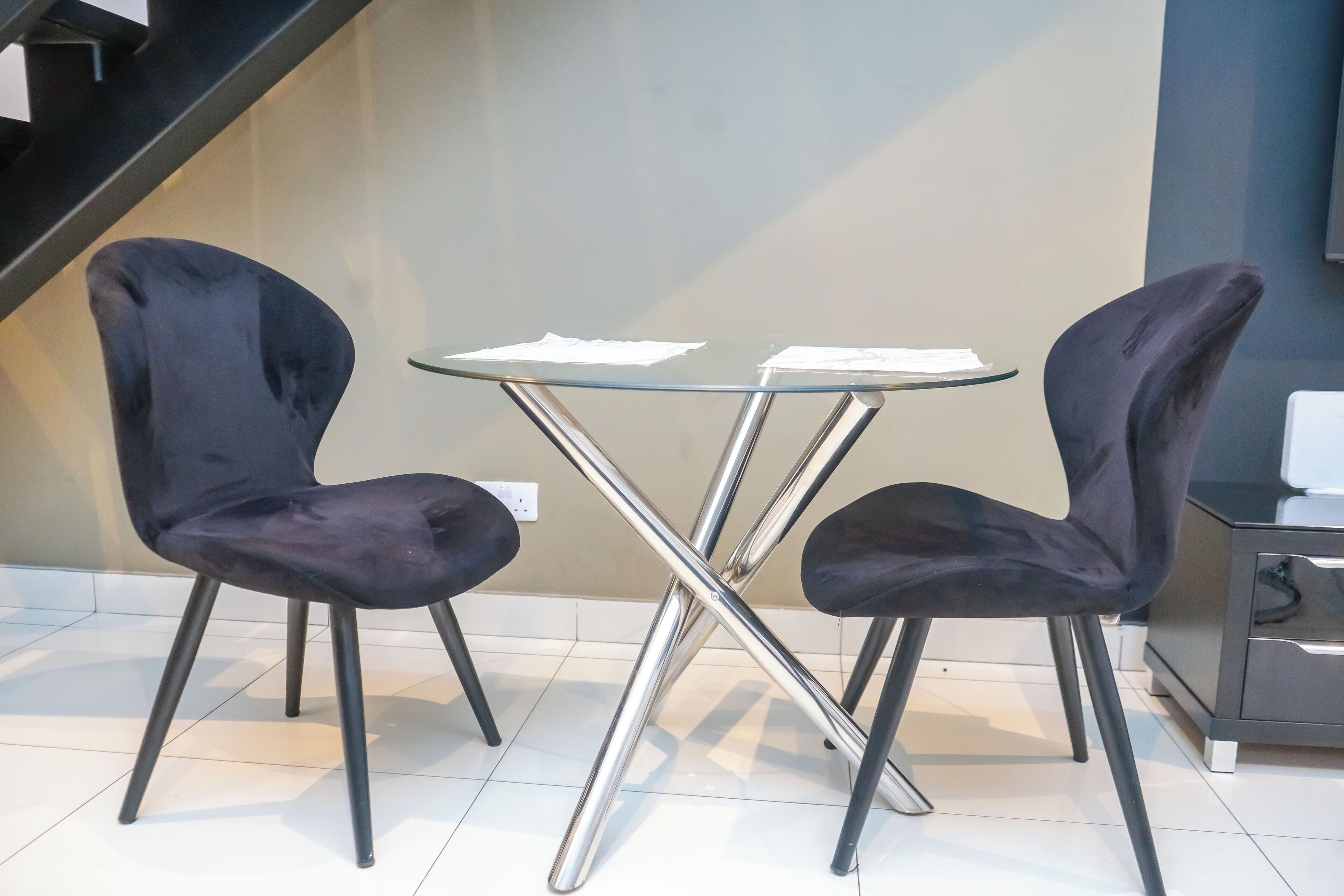 Una habitación moderna con una mesa de vidrio y dos sillas negras, una escalera al fondo y un armario a la derecha.
