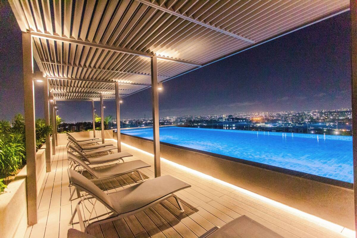 A rooftop pool area with lounge chairs, a view of the cityscape at night, and a covered pergola structure.