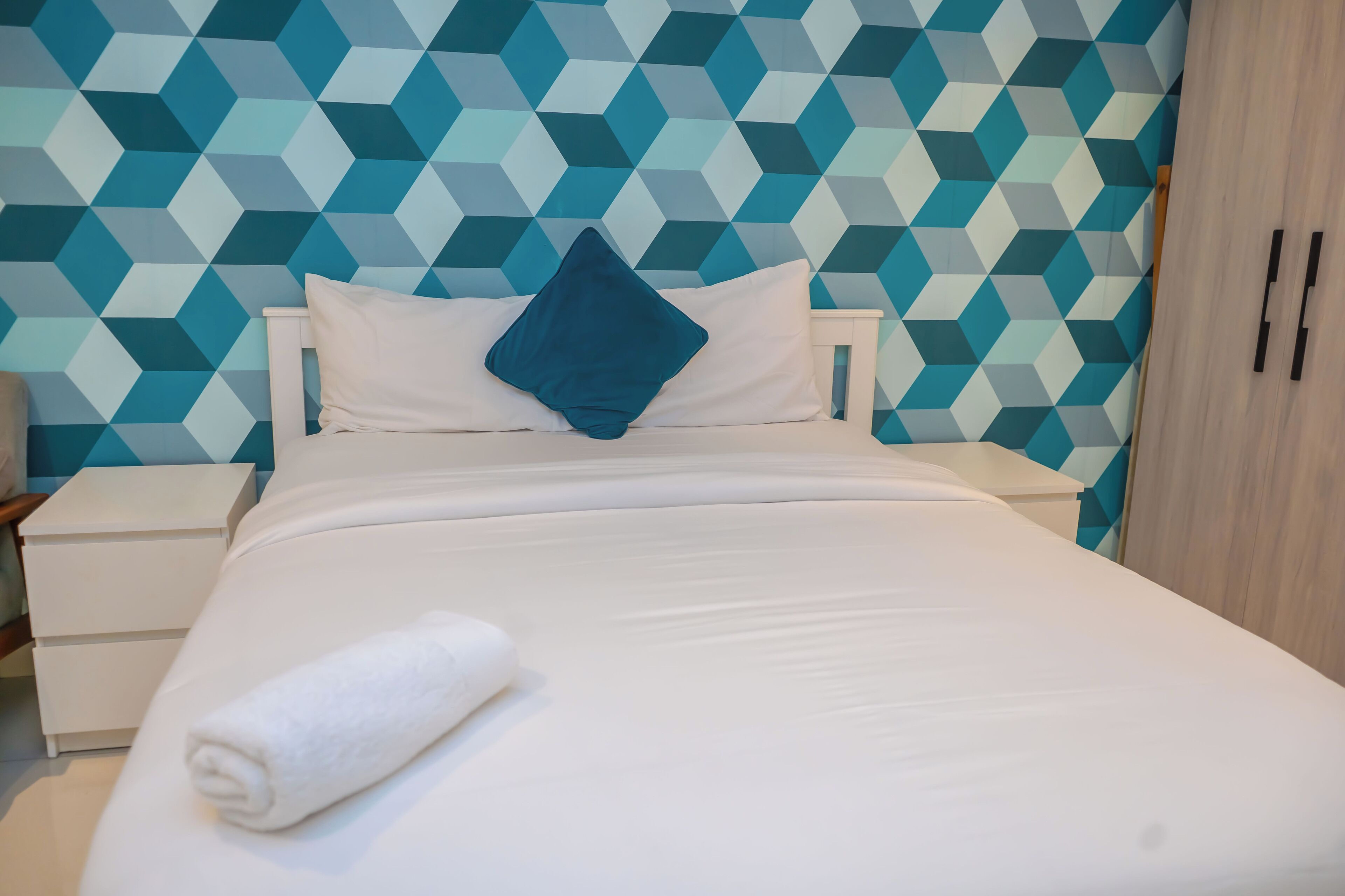 Un lit bien fait, avec une couette blanche et un oreiller bleu, une table de chevet blanche et un mur orné d’un motif géométrique.