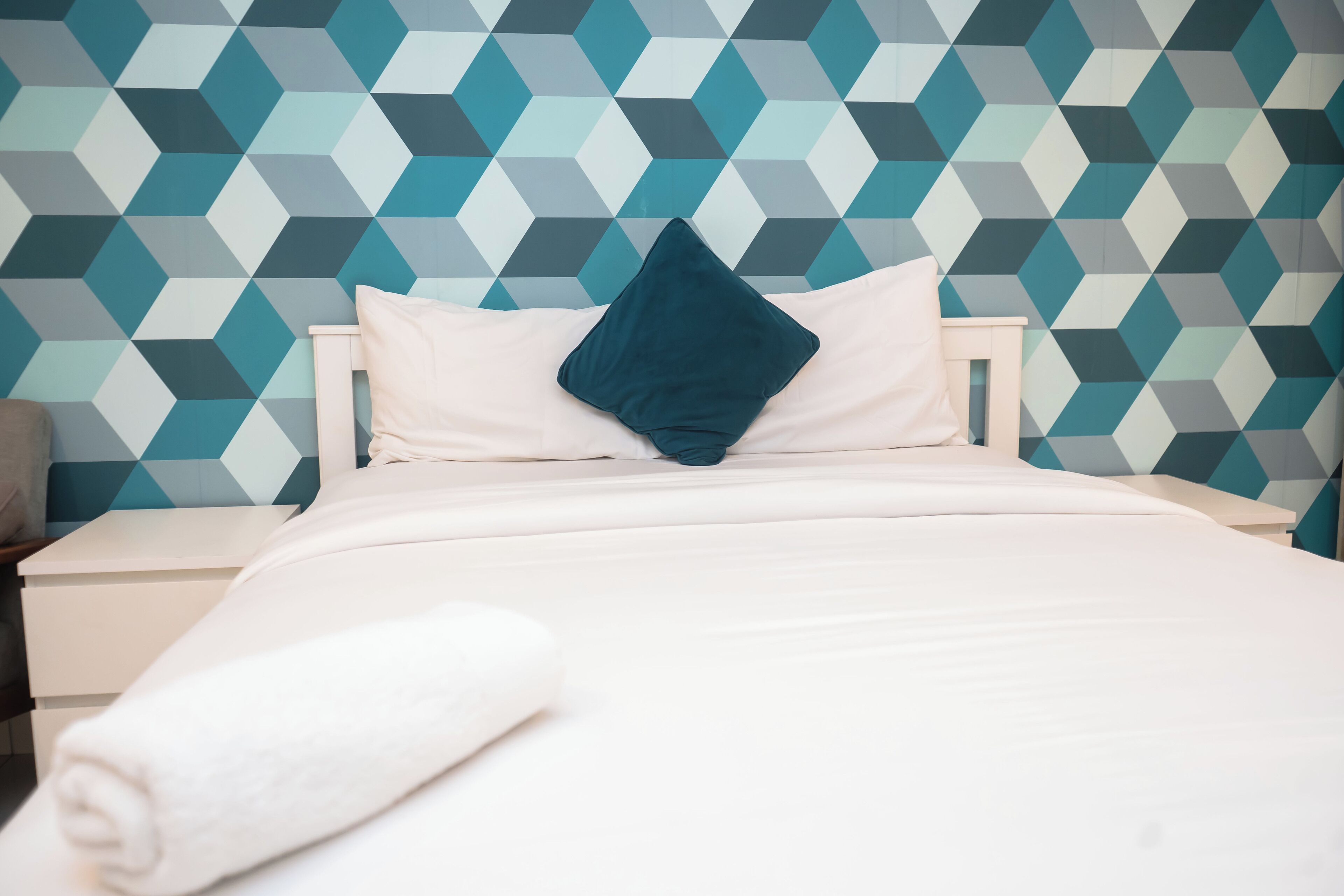 Una cama bien hecha con una colcha blanca y un almohadón turquesa, frente a una pared con diseño geométrico.