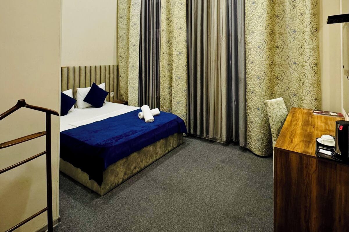 A hotel room with a bed, a desk, and a chair. There is a window with curtains and an air conditioning unit on the wall.