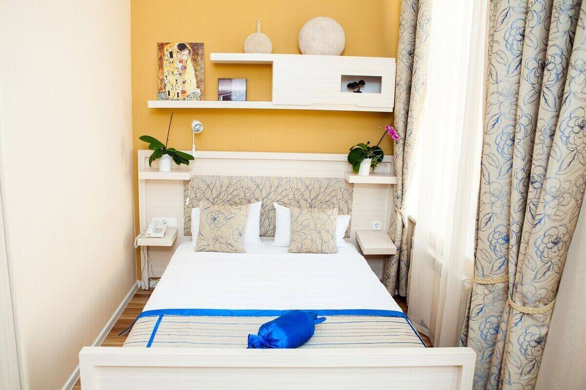 A hotel room with a bed, bedside tables, a wall-mounted shelf with decorative items, and a window with curtains.