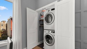 A laundry room with two stacked front-loading washing machines, a drying rack, and a small shelf with laundry supplies.
