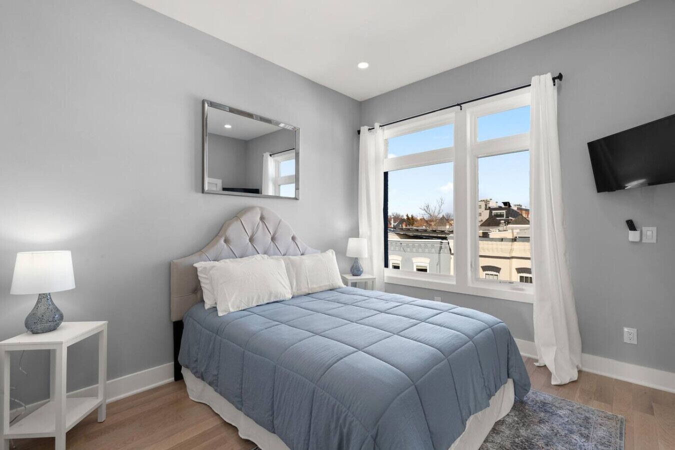 A bedroom with a bed, a nightstand, a lamp, a mirror, a window with curtains, and a TV on the wall.