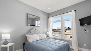 A bedroom with a bed, a nightstand, a lamp, a mirror, a window with curtains, and a TV on the wall.