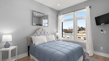 A bedroom with a bed, a nightstand, a lamp, a mirror, a window with curtains, and a TV on the wall.