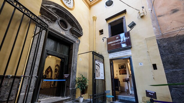 A narrow street with a building entrance, a potted plant, and a chair outside. - Vico Santa Luciella 20 (Naples)