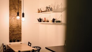 A dining area with a wooden table, chairs, and pendant lights. There is a stone wall behind the table and shelves above holding kitchen items. - Vico Santa Luciella 20 (Naples)