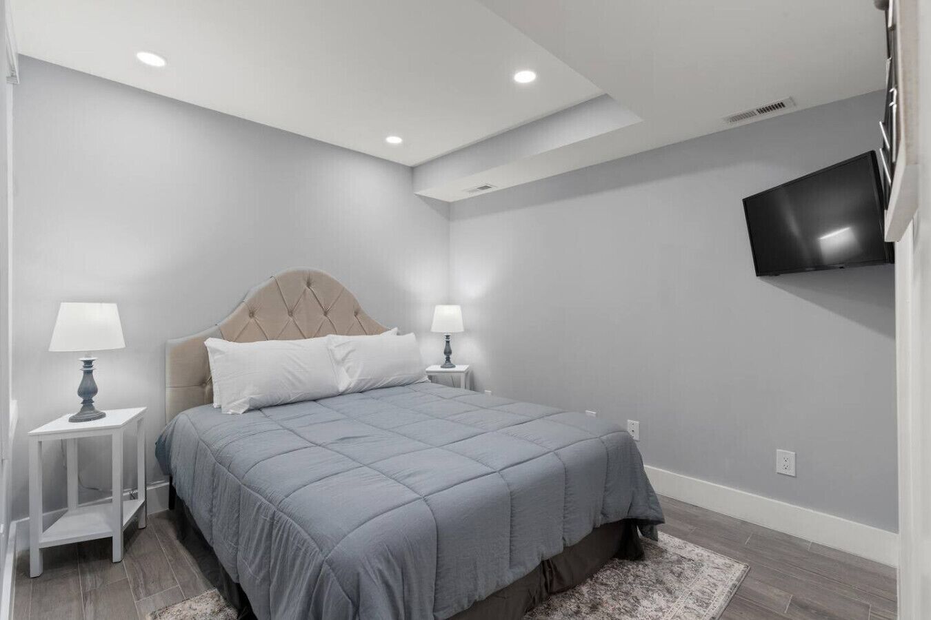 Ein modernes Schlafzimmer mit einem Bett, zwei Nachttischen mit Lampen, einem an der Wand befestigten Fernseher und Einbauleuchten.