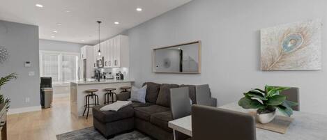 A modern living room with a sectional sofa, a dining table with chairs, a kitchen in the background, and a large abstract painting on the wall.