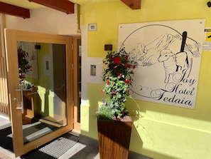 Ingresso di un hotel con porta in legno, tappetino d'entrata, una pianta in vaso con fiori rossi e un cartello che riporta 'Hotel Fediaia' con un'illustrazione di una pecora.