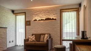 A modern living room with a stone wall, a brown sofa, a wooden coffee table, and a large window. - Room 'Fedaia Joy Bnb Suite' with Mountain View, Balcony and Wi-Fi (Canazei)