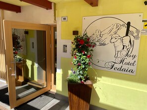 A hotel entrance with a wooden door, a welcome mat, a potted plant with red flowers, and a wall sign for 'Hotel Fediaia' with a sheep illustration.