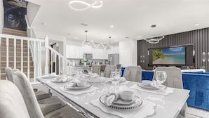 Un espace repas moderne avec une longue table dressée pour six personnes, une cuisine en arrière-plan et un téléviseur fixé au mur.