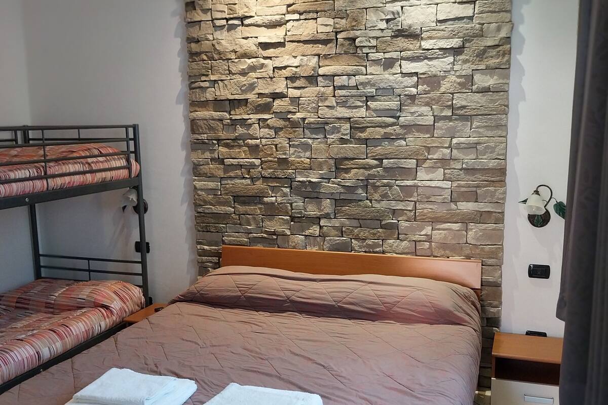A bedroom with a bunk bed, a single bed, a stone wall, a bedside table, a lamp, and a tiled floor.