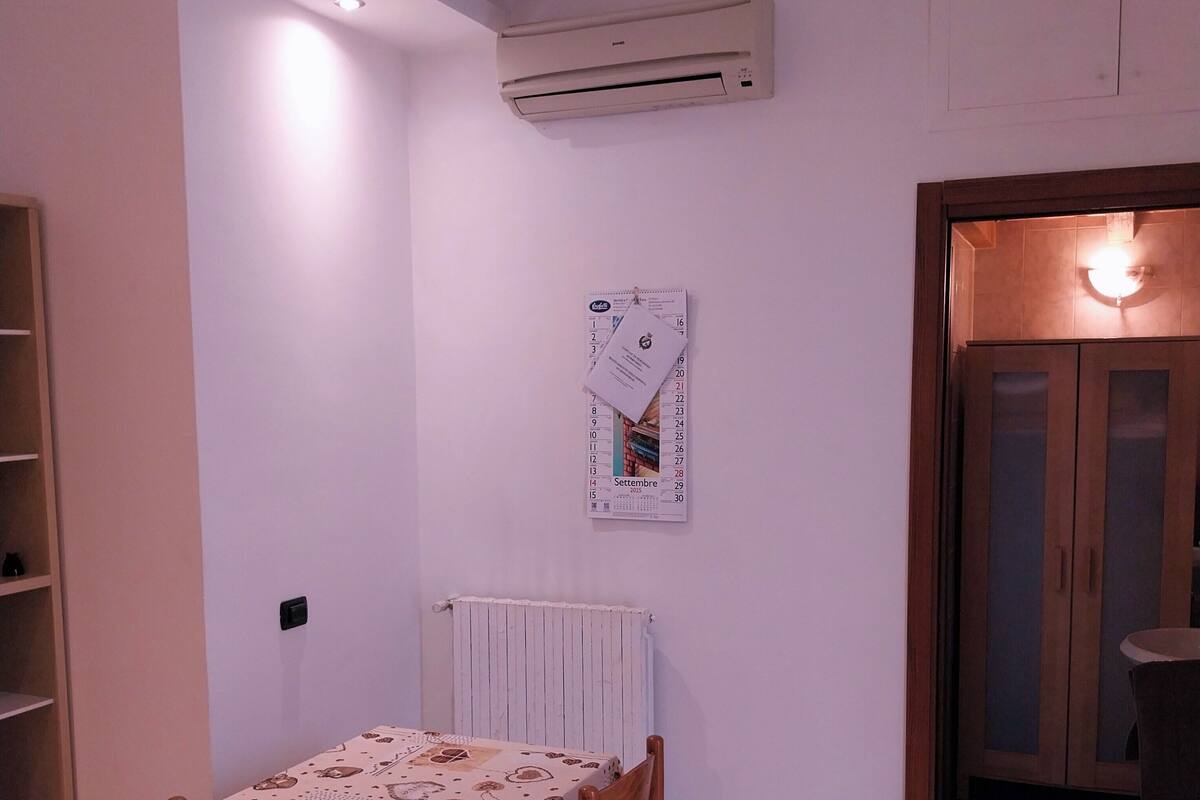 A dining area with a table covered by a patterned tablecloth, wooden chairs, an air conditioning unit, a calendar on the wall, and a door leading to another room.