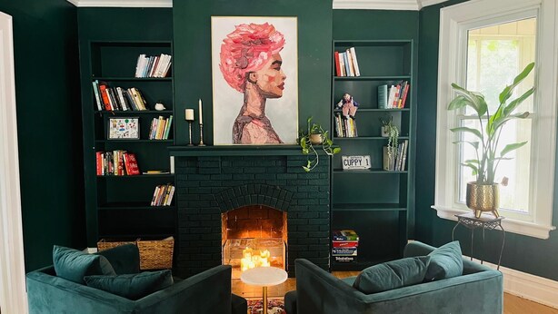 Een woonkamer met een open haard, donkergroene wanden en boekenkasten vol boeken.