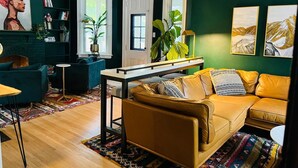 Een moderne woonkamer met een bank, salontafel en een open haard. Er staan boekenkasten en een groot raam met planten.
