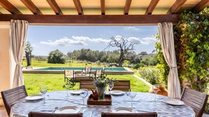 A dining area with a table set for four, a view of a swimming pool, and a pergola with curtains. - Countryside finca in Mallorca with Private Pool, Tradition and Nature (Illes Balears)