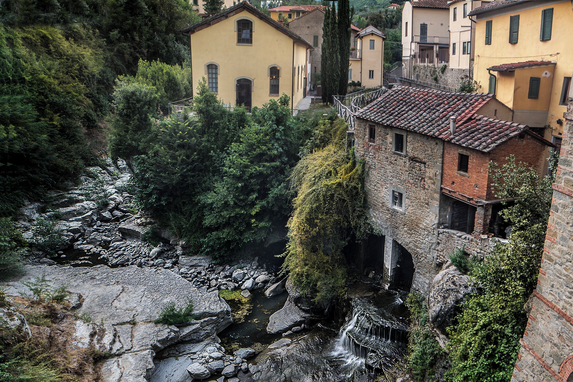 A stone building with a small waterfall, surrounded by lush greenery and traditional yellow houses.