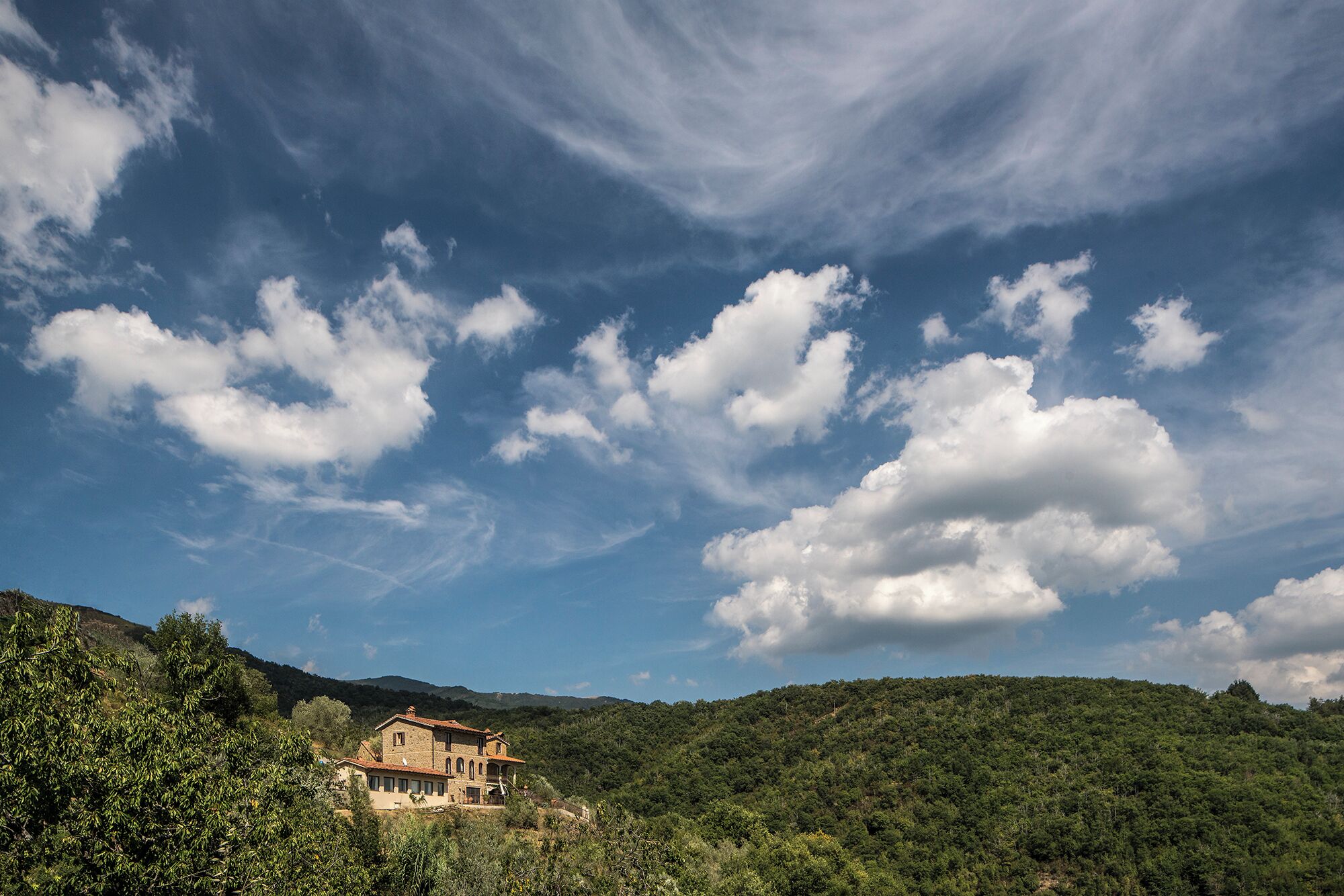A rustic stone building with a tiled roof, surrounded by lush green hills and a clear blue sky with scattered clouds.