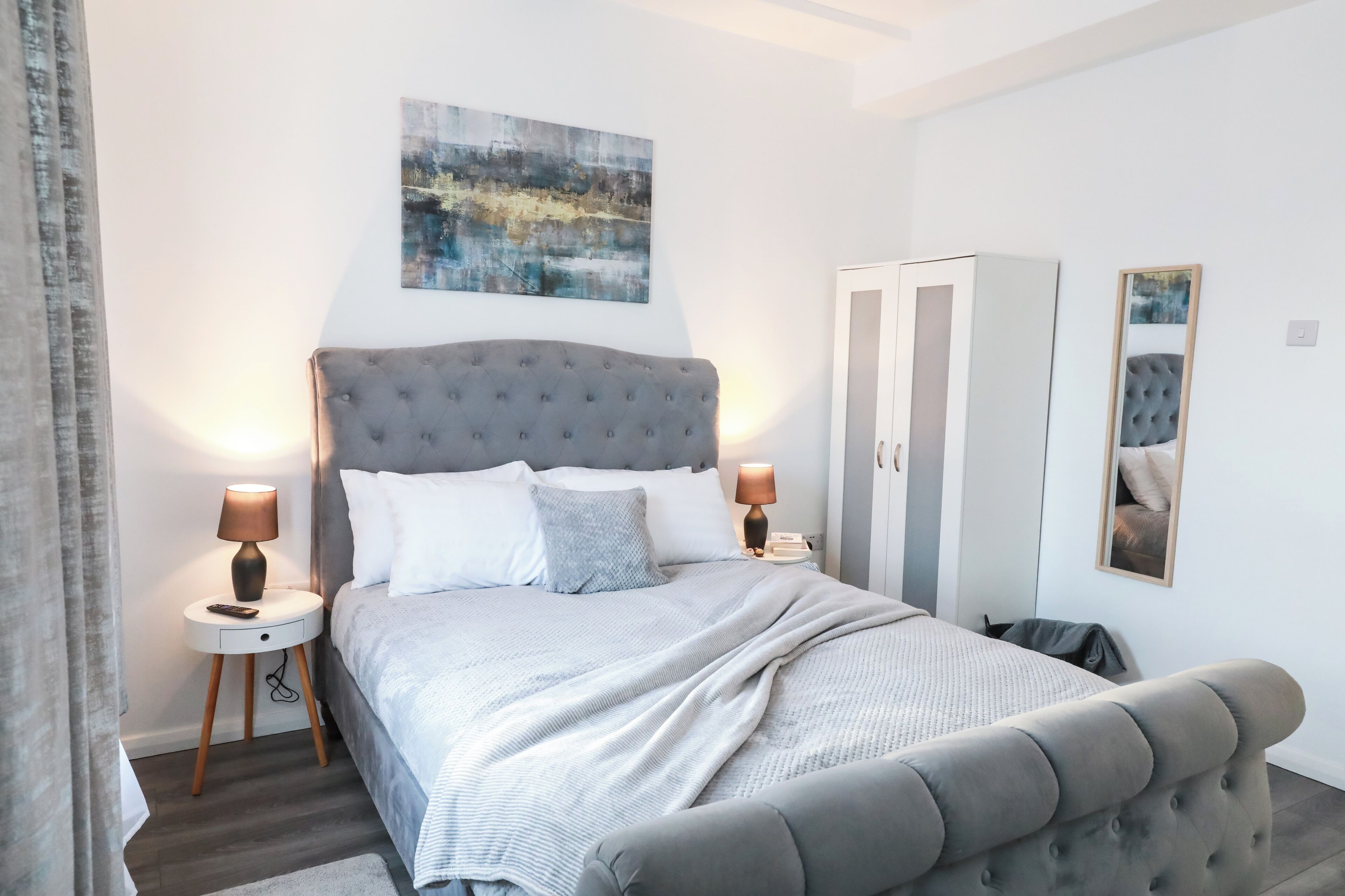 Ein Schlafzimmer mit einem gepolsterten Kopfteil, einer grauen Bettdecke, zwei Nachttischlampen, einem weißen Nachttisch, einem Kleiderschrank und einem gerahmten Spiegel an der Wand.