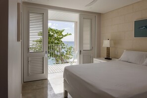 Een hotelkamer met een bed, nachtkastje, lamp en uitzicht op de oceaan door open luiken.