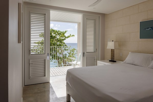A hotel room with a bed, a nightstand, a lamp, and a view of the ocean through open shutters.