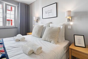 A hotel room with a neatly made bed, two wall-mounted lamps, a framed quote on the wall, and a window with curtains.