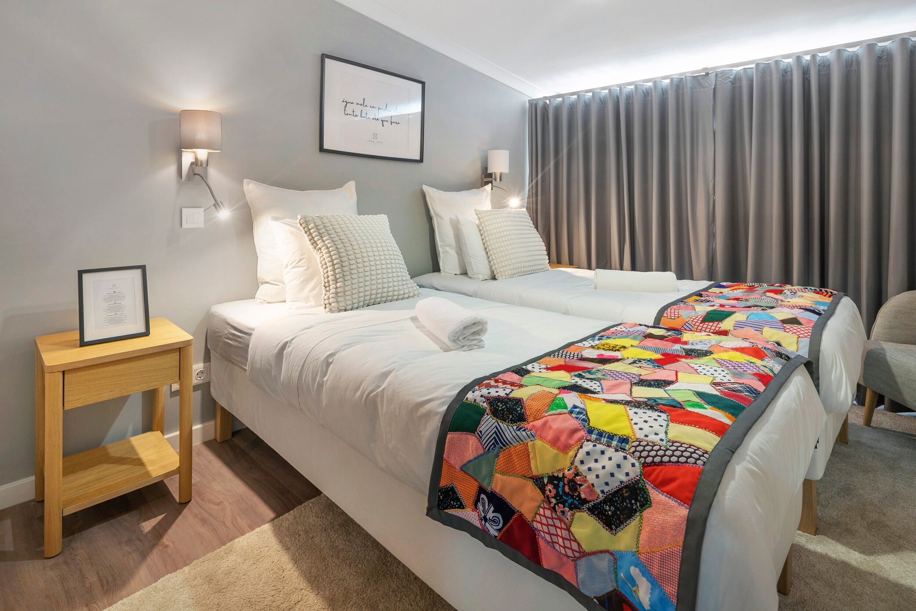 A double bed with a colorful quilt, a bedside table with a framed certificate, and a wall-mounted light fixture.
