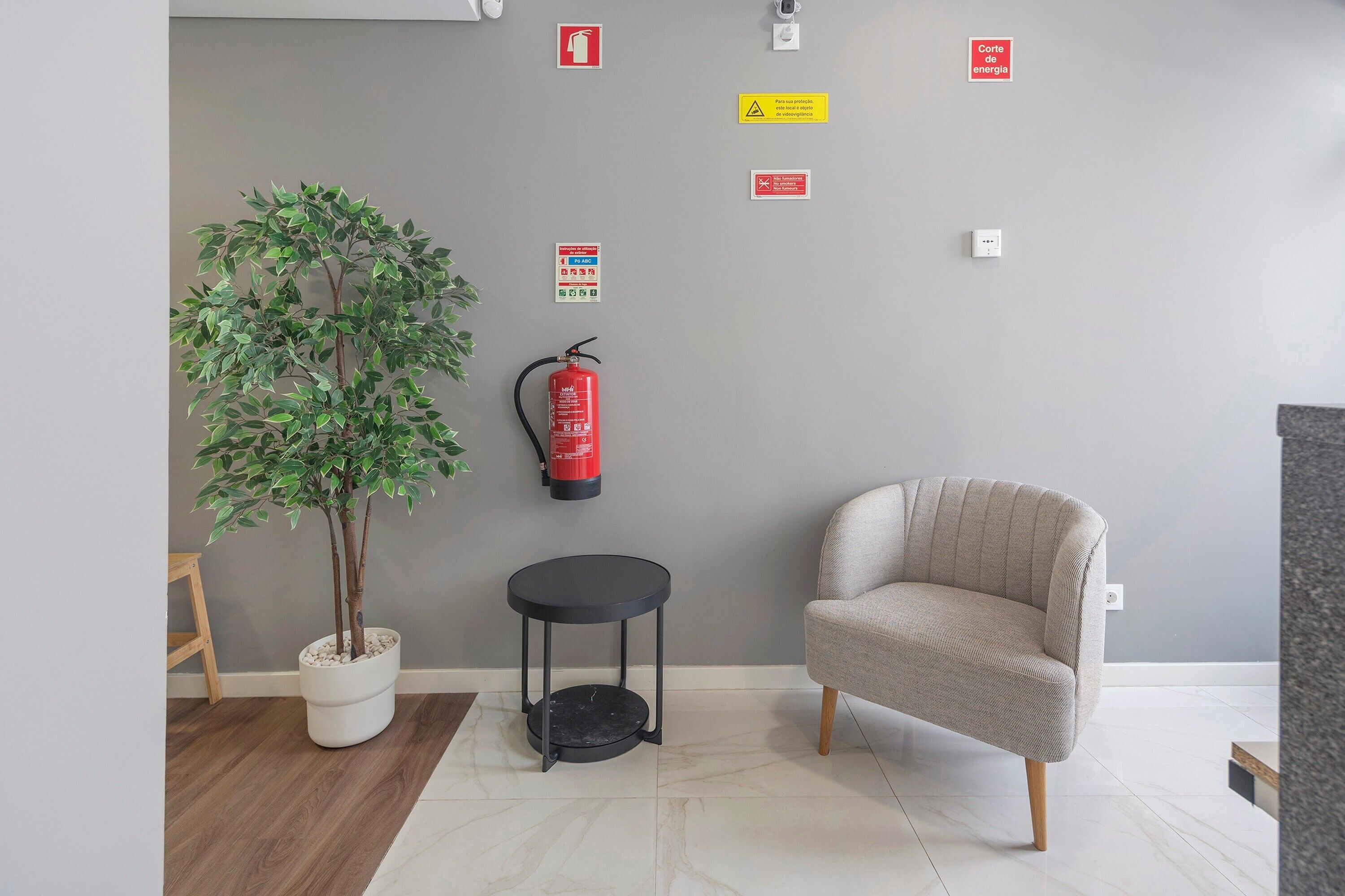 Een kamer met een potplant, een ronde zwarte tafel, een beige fauteuil, een brandblusser en diverse veiligheidspictogrammen aan de muur.