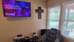 A living room with a television displaying a connectivity issue message, a cross on the wall, a window with blinds, a grey armchair, and a small table with items on it. - Private cabin for two
National Forest and outdoor fireplace 
Near Hot Springs (Glenwood)