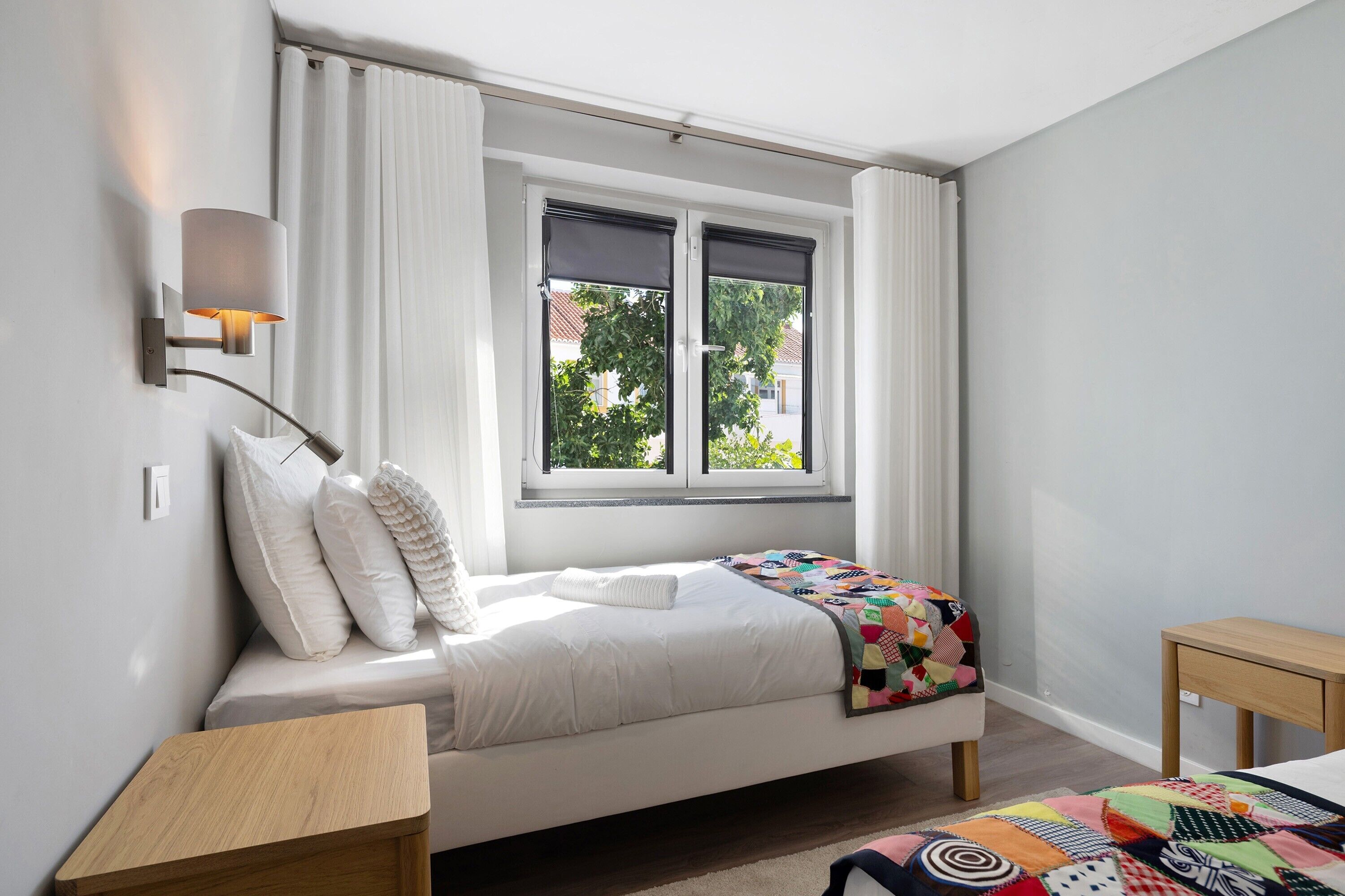 Ein Schlafzimmer mit Bett, Nachttisch, einem Fenster mit Vorhängen und einer Wandlampe.