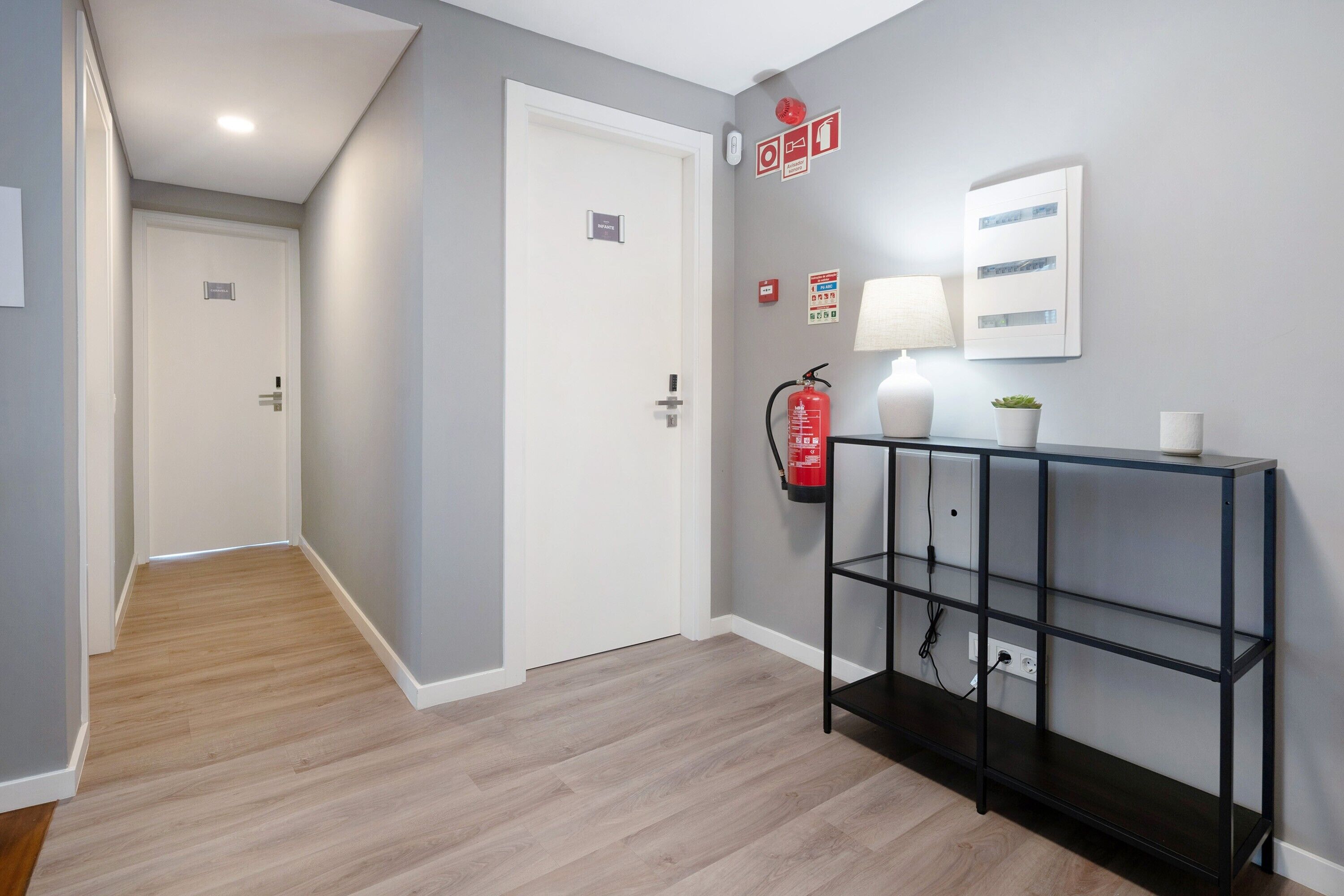 A hallway with a white door, a black metal shelf with a lamp, and a fire extinguisher on the wall.