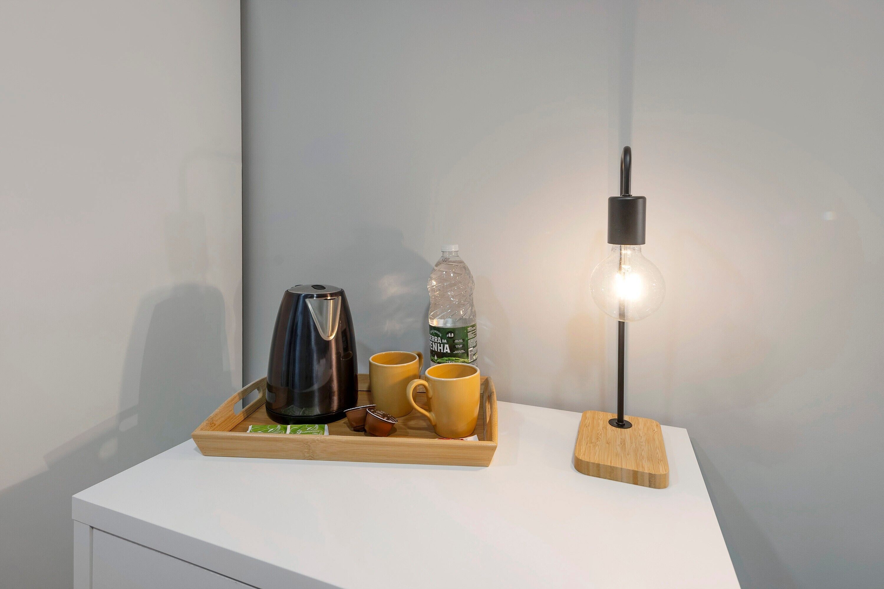 A white table with a tray containing a black coffee pot, two yellow cups, a bottle of water, and a wooden lamp.