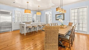A modern kitchen with white cabinetry, a central island, and bar stools. There is a dining area with a wooden table and chairs. - NEW 2 Blocks to Camp WaterColor with LSV, Bikes, and more! (Santa Rosa Beach)