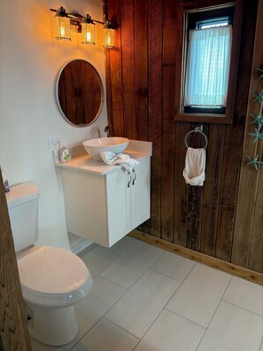 A bathroom with a white toilet, a round mirror, a white sink with a white towel, and wooden paneling on the walls.