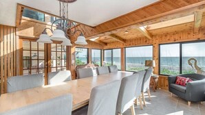 Um quarto espaçoso com uma grande mesa de jantar de madeira, rodeada de cadeiras, amplas janelas com vista para o mar e uma área de estar aconchegante com um sofá.