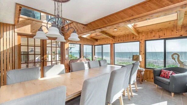 A spacious room with a large wooden dining table surrounded by chairs, large windows offering a view of the sea, and a cozy seating area with a sofa.