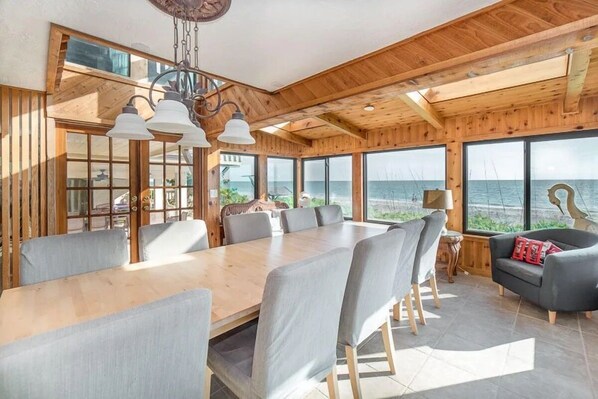 A spacious room with a large wooden dining table surrounded by chairs, large windows offering a view of the sea, and a cozy seating area with a sofa.