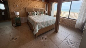 A bedroom with a wooden ceiling, a bed with a patterned bedspread, a nightstand with a lamp, and a view of the beach through large windows.
