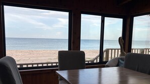 Quarto com uma janela grande com vista para a praia. Há uma mesa de madeira e duas cadeiras.